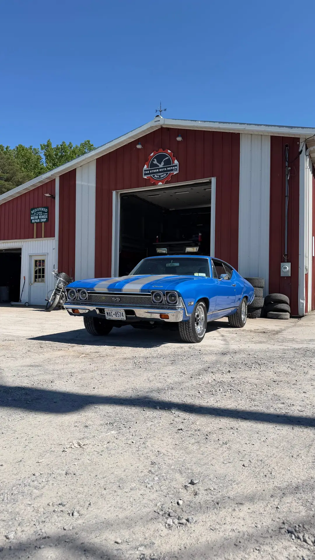 A blue classic muscle car with white racing stripes parked outside an open bay at The Other Guys Repair